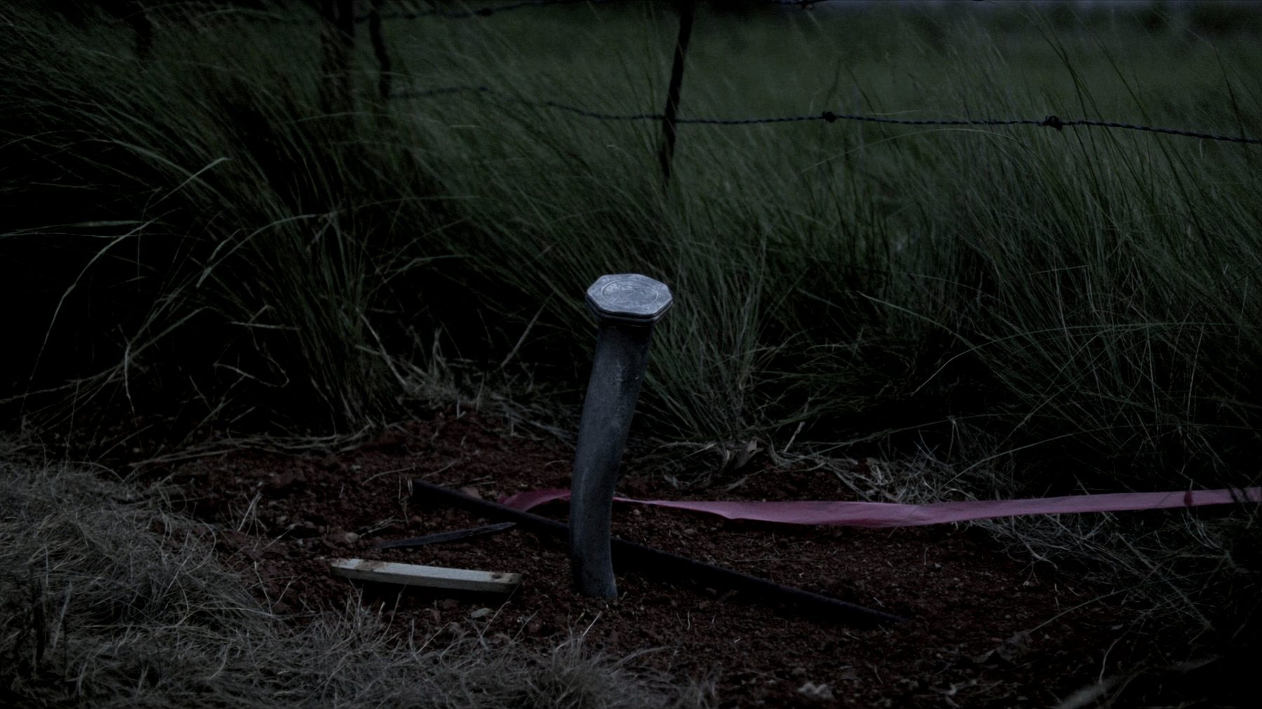 A rusted iron survey monument with a small stamped cap, driven into hard clay at the edge of a pasture fence line, with dry grass and a strand of barbed wire behind it at dusk.